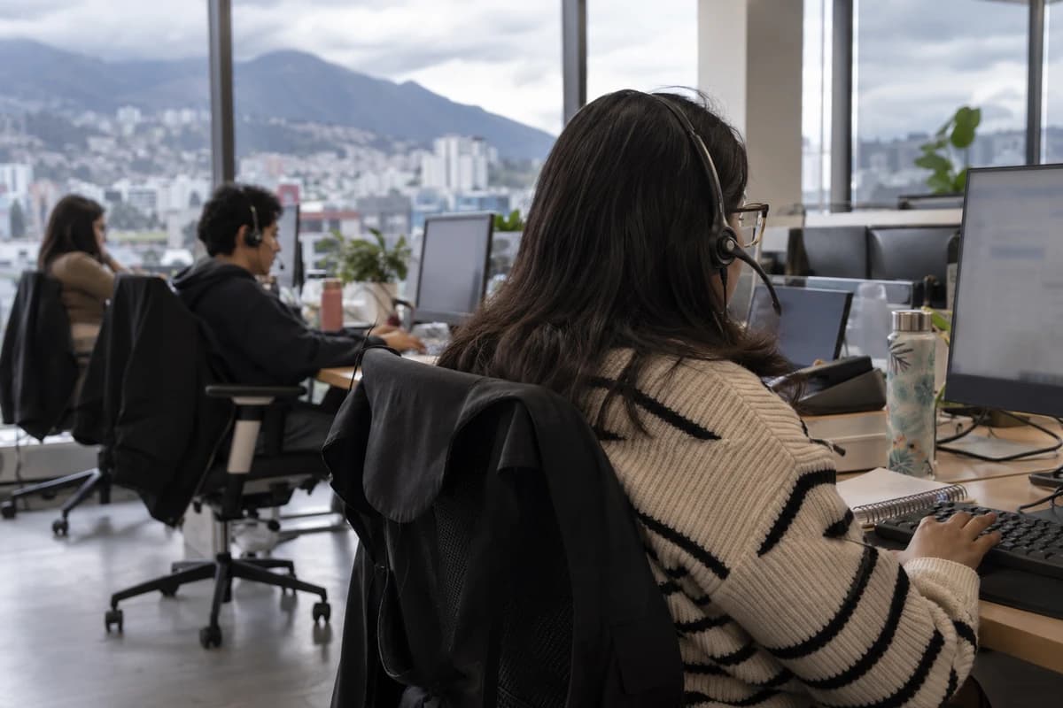Oficina de atención al cliente en Quito: una agente de espaldas, con auricular, escribe en su teclado; al fondo, dos compañeros trabajan en sus puestos frente a un ventanal con la ciudad y el Pichincha.